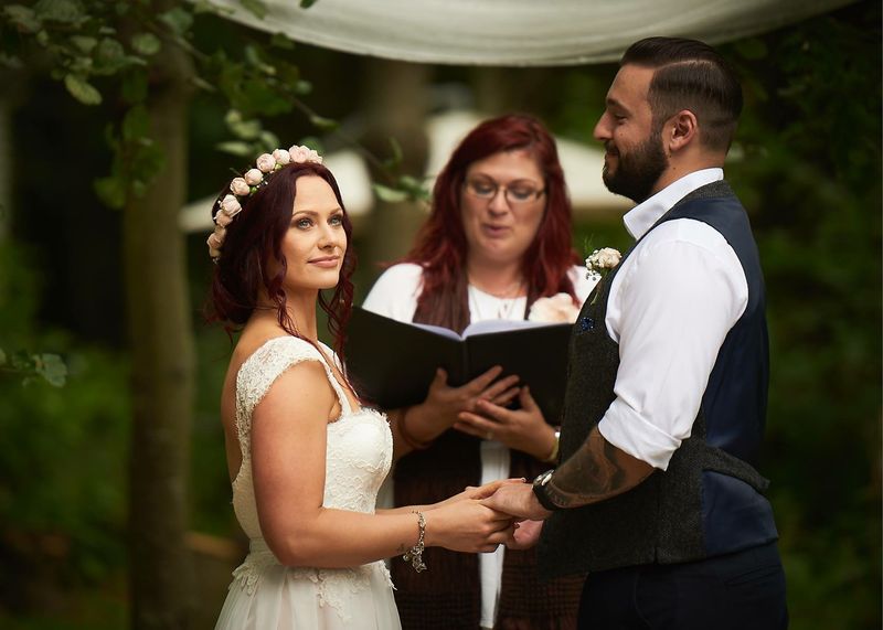 Roxanne reading from her ceremony book as a bride and groom hold hands under a woodland canopy